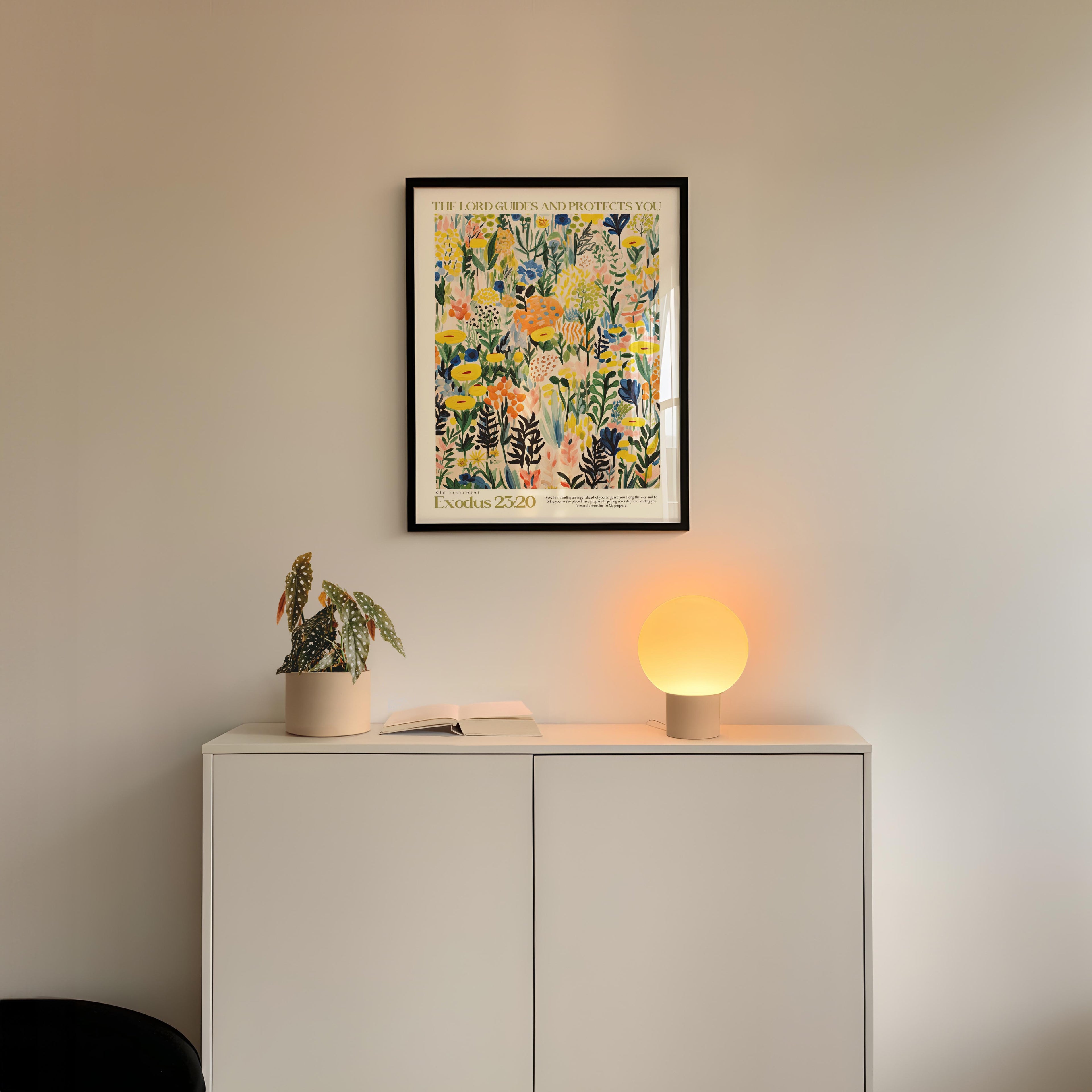 a white cabinet with a plant and a lamp on top, and a framed poster hanging on the wall above the cabinet.