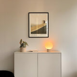 a minimalist interior design with a white cabinet, a potted plant, and a lamp on a shelf. Above the cabinet, there is a framed poster of a desert landscape.
