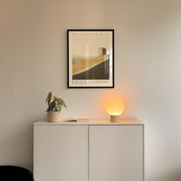 a minimalist interior design with a white cabinet, a potted plant, and a lamp on a shelf. Above the cabinet, there is a framed poster of a desert landscape.