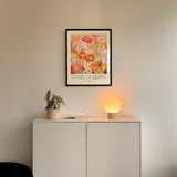 a white cabinet with a potted plant and a lamp on top, set against a white wall. Above the cabinet, there is a framed poster with a colorful floral design.