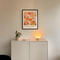 a white cabinet with a potted plant and a lamp on top, set against a white wall. Above the cabinet, there is a framed poster with a colorful floral design.