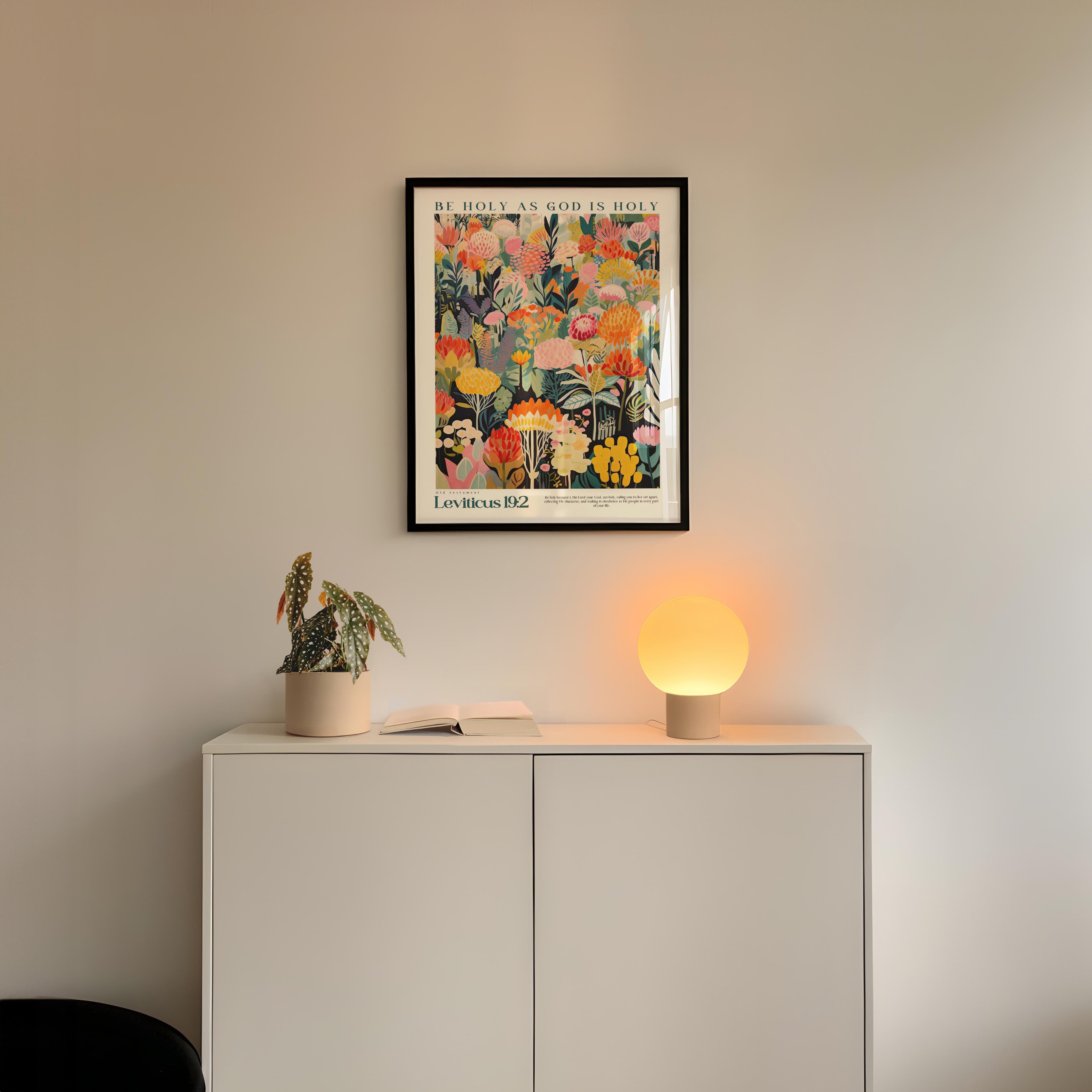 a white cabinet with a potted plant and a lamp on top, set against a white wall. Above the cabinet, there is a framed poster with a colorful floral design.