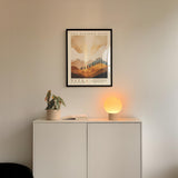 a minimalist interior design with a framed poster of a landscape, a potted plant, and a small lamp on a white cabinet.