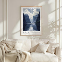 a framed poster of a serene mountain landscape with a lake, hanging on a wall above a white couch with a blanket draped over it.