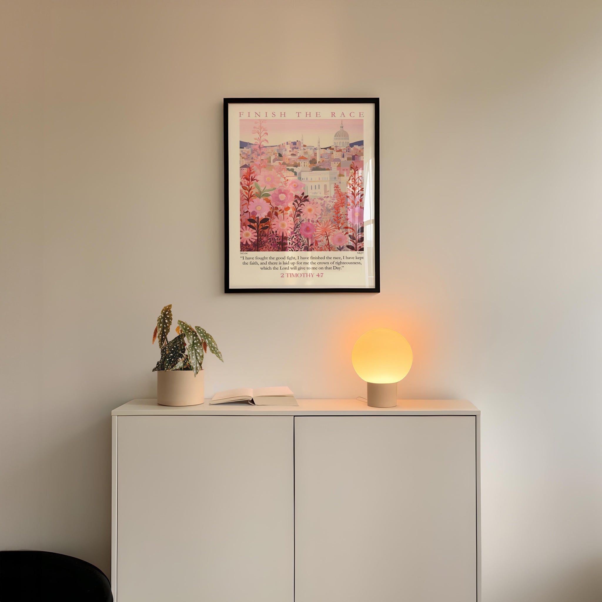 a white cabinet with a potted plant and a lamp on top, set against a white wall. Above the cabinet, there is a framed poster with a pink and white floral design.
