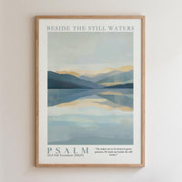 The image is a framed poster with a serene landscape depicting a calm body of water surrounded by mountains. The text "BESIDE THE STILL WATERS" is written at the top of the poster, and the phrase "PSALM" is written at the bottom. The poster also includes a Bible verse from the Old Testament (NIV) that reads, "He makes us to lie down in green pastures, leads us beside the still waters."