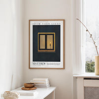 a framed poster of a lit candle on a wall, with a window and a white table with books and a bowl on it in the foreground.