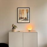 a minimalist interior design with a framed poster of a religious scene hanging on a white wall above a white cabinet. The cabinet has a plant and a lamp on it, creating a cozy and serene atmosphere.