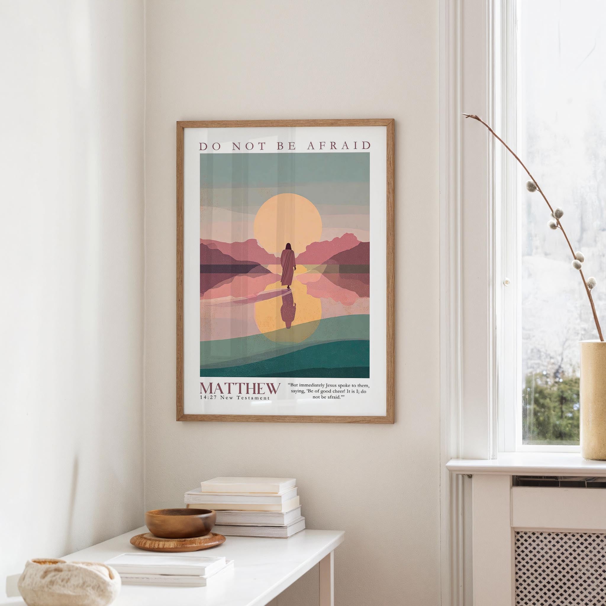 a framed poster of a sunset landscape hanging on a wall, with a stack of books and a vase on a table below it.