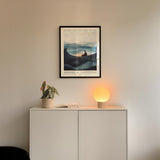 a minimalist interior design with a framed poster of a landscape, a potted plant, and a small lamp on a white cabinet.