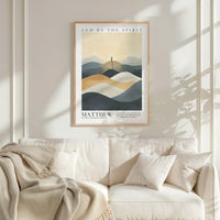 a framed poster of a mountain landscape with a person standing on top, hanging on a wall above a white couch with a blanket draped over it.