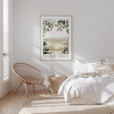 A bedroom with a bed, a woven chair, and a framed poster of a landscape on the wall.