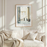 a framed poster of a classical architectural scene hanging on a white wall above a white couch with a blanket draped over it.