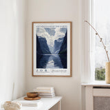 a framed poster of a mountainous landscape with a lake, hanging on a wall in a room with a white table and a vase.