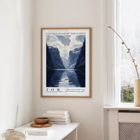 a framed poster of a mountainous landscape with a lake, hanging on a wall in a room with a white table and a vase.