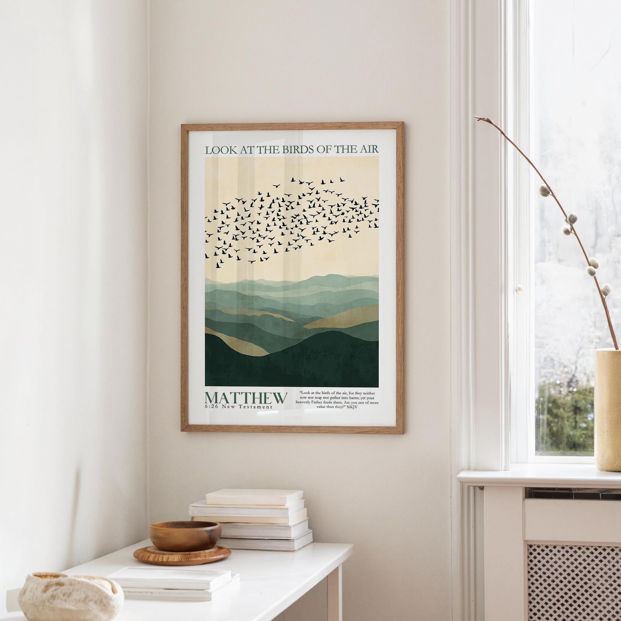 a framed poster of a flock of birds flying over a mountainous landscape, hanging on a wall in a room with a white table and a window.