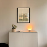 a minimalist interior design with a framed poster of a tree and mountains on the wall above a white cabinet with a plant and a lamp on top.