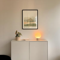 a minimalist interior design with a framed poster of a tree and mountains on the wall above a white cabinet with a plant and a lamp on top.