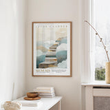 A framed poster of a staircase leading to clouds hangs on a wall in a room with a white table and a vase of flowers.