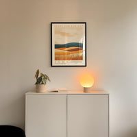 a minimalist interior design with a framed landscape painting hanging on a white wall above a white cabinet. The cabinet has a plant and a lamp on it, creating a cozy and inviting atmosphere.