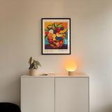a white cabinet with a potted plant and a lamp on top, and a framed poster of a fruit bowl hanging on the wall above the cabinet.