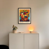 a white cabinet with a potted plant and a lamp on top, and a framed poster of a fruit bowl hanging on the wall above the cabinet.