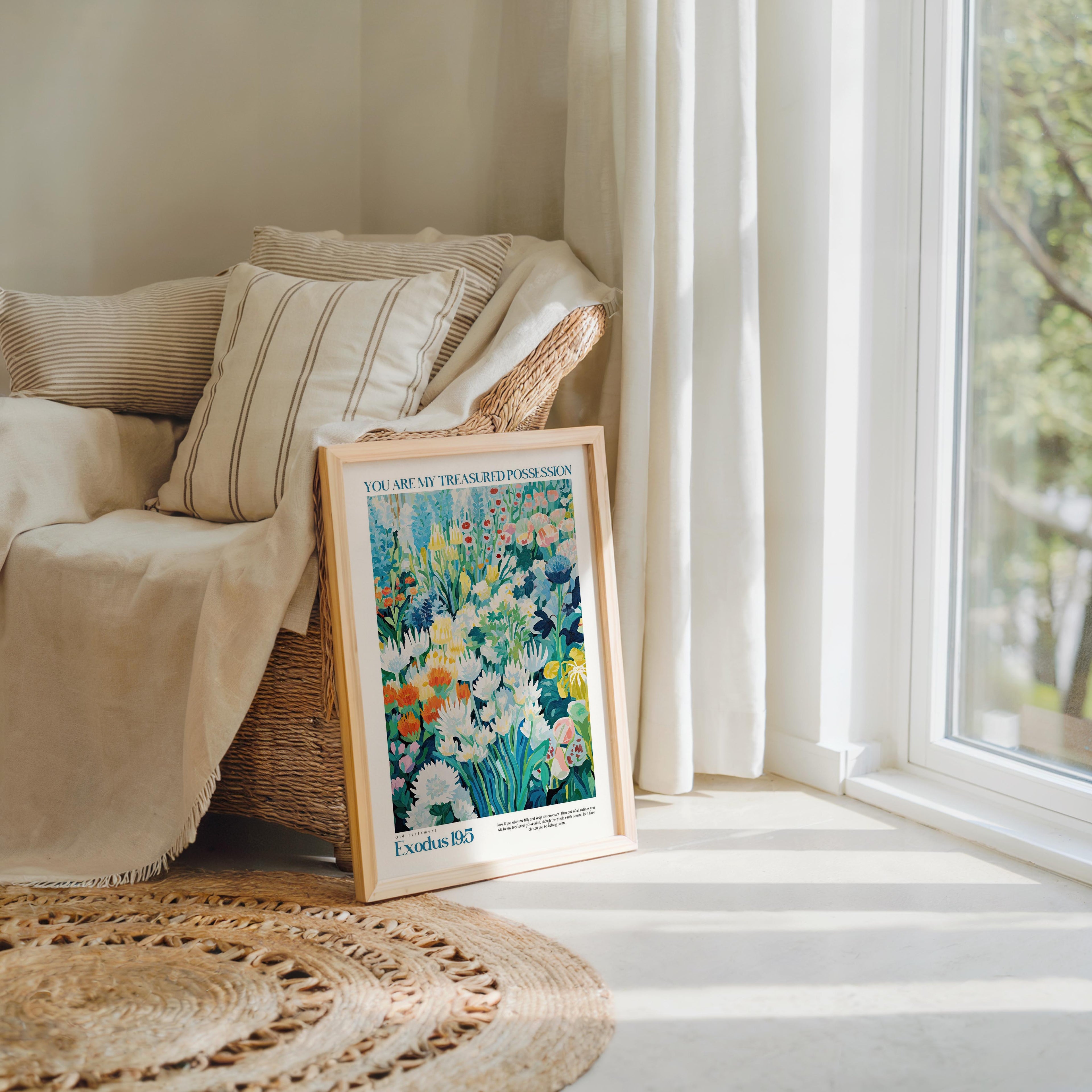 A framed poster of a colorful floral painting is propped up against a beige armchair in a cozy living room, with a large window and a woven rug visible in the background.