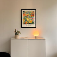 a white cabinet with a potted plant and a lamp on top, set against a white wall. Above the cabinet, there is a framed poster with a floral design.