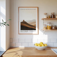 a kitchen counter with a bowl of lemons, a framed poster of a desert landscape, and a shelf with jars and containers.