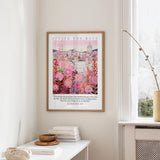 a framed poster of a cityscape with pink flowers hanging on a wall, next to a window and a small table with books and a bowl.