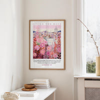 a framed poster of a cityscape with pink flowers hanging on a wall, next to a window and a small table with books and a bowl.