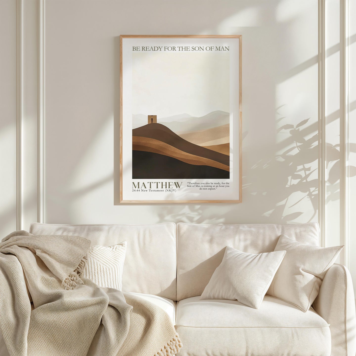 a framed poster of a mountain landscape with the text "BE READY FOR THE SON OF MAN" above it, hanging on a white wall above a white couch with a blanket draped over it.