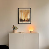 a minimalist interior design with a framed poster of silhouetted figures hanging on a wall above a white cabinet. The cabinet has a plant and a lamp on top of it, creating a cozy and inviting atmosphere.