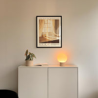 a minimalist interior design with a framed poster of a window and curtains, a small potted plant, and a white cabinet with a lamp on top.