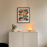 a white cabinet with a potted plant and a lamp on top, and a framed poster of a colorful abstract design hanging on the wall above the cabinet.