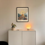 a white cabinet with a potted plant and a lamp on top, and a framed poster of a religious scene hanging on the wall above it.