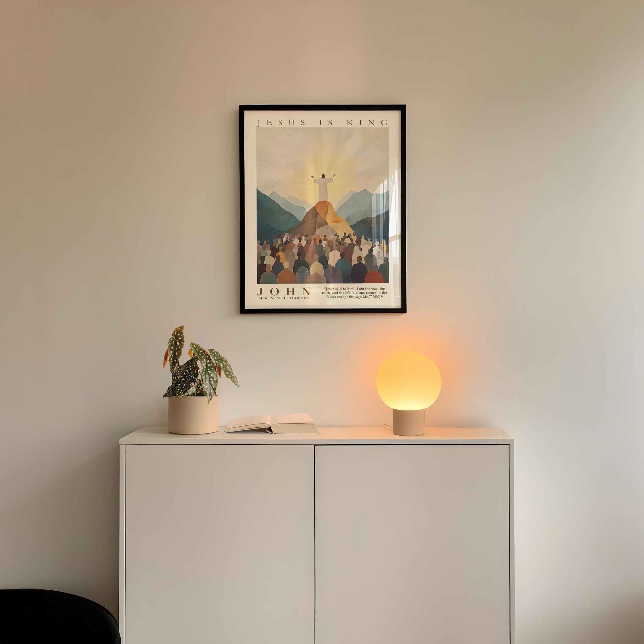 a white cabinet with a potted plant and a lamp on top, and a framed poster of a religious scene hanging on the wall above it.