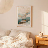 A framed poster of a staircase leading to clouds hangs on a wall above a bed with striped pillows.