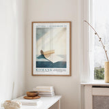 a framed poster of a blue and white abstract design hanging on a wall, with a white table below it holding a stack of books and a small vase.