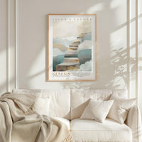 A framed poster of a staircase leading up to a cloud-filled sky hangs on a white wall above a beige couch with a blanket draped over it.