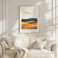 a framed poster of a landscape with mountains and a field, hanging on a wall above a white couch with a blanket draped over it.