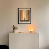 a minimalist interior design with a framed poster of a building, a potted plant, and a small lamp on a white cabinet.