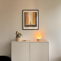 a minimalist interior design with a framed poster of a building, a potted plant, and a small lamp on a white cabinet.