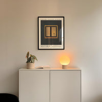 a minimalist interior design with a framed poster of a window, a potted plant, and a small lamp on a white cabinet.