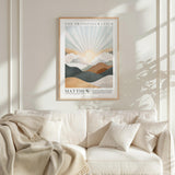a framed poster of a mountain landscape with a sunburst effect, hanging on a white wall above a white couch with a blanket draped over it.