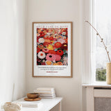 a framed poster of a vibrant floral arrangement hanging on a wall, with a stack of books and a small vase on a white table below it.