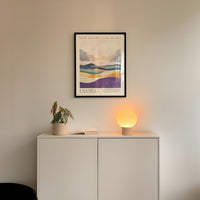 a minimalist interior design with a framed landscape poster on the wall above a white cabinet, which has a plant and a lamp on top of it.