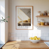 A framed poster of a basket of bread and a bowl of lemons hangs on a white tiled wall above a wooden countertop with a bowl of lemons.