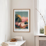 a framed poster of a landscape with mountains and trees, hanging on a wall above a white table with a stack of books and a small vase.