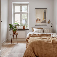 a cozy bedroom with a large bed, a wooden nightstand, and a potted plant. The room has a neutral color scheme with white walls and a wooden floor. A framed poster is hanging on the wall above the bed, and a window with a plant on the sill is visible.